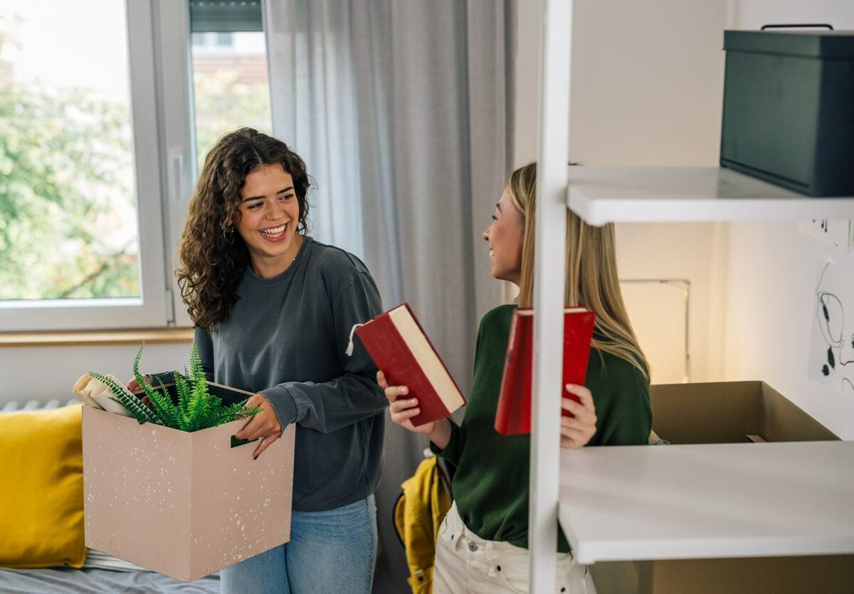 Two students hold boxes and book as they talk and laugh in a college dorm room