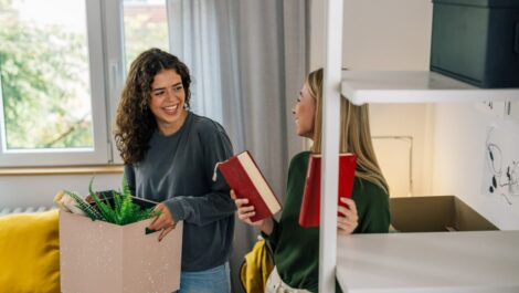 Two students hold boxes and book as they talk and laugh in a college dorm room
