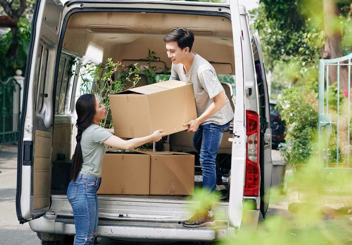 A man stands in a van and lowers a cardboard box to a woman standing on the ground