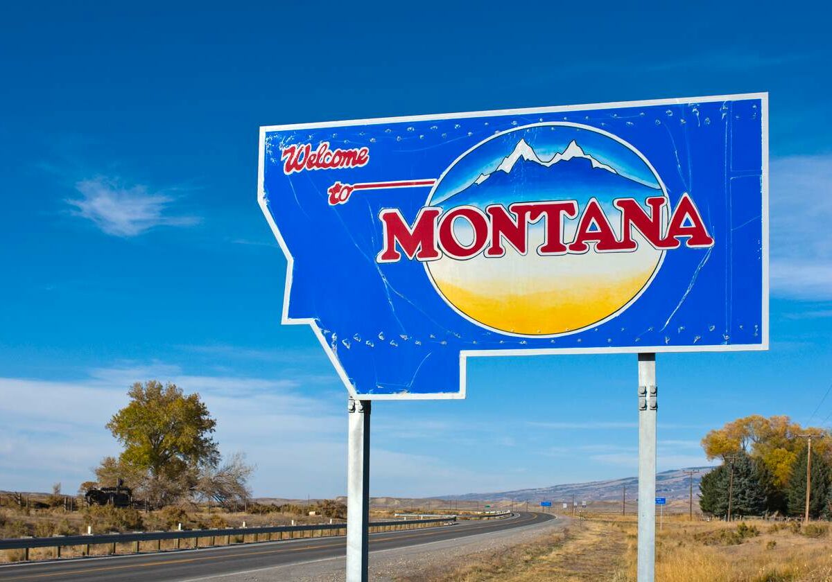 A blue sign in the shape of Montana reads 'Welcome to Montana.' In the background is a highway.