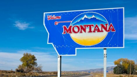 A blue sign in the shape of Montana reads 'Welcome to Montana.' In the background is a highway.