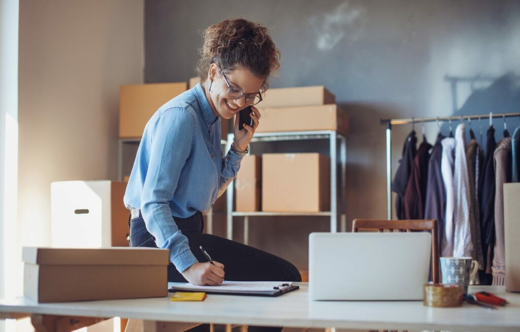 A woman talking on a cell phone takes notes on a clipboard. Racks of clothing and boxes sit in the background. 