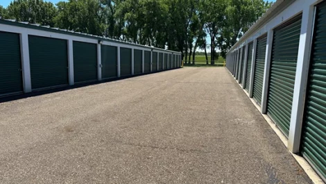 Storage units on both sides of a paved road with surrounding trees.