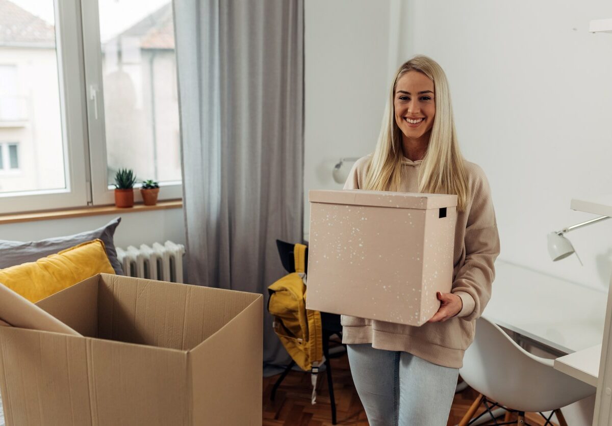 A college student smiles as she unpacks boxes of her items in her dorm room.