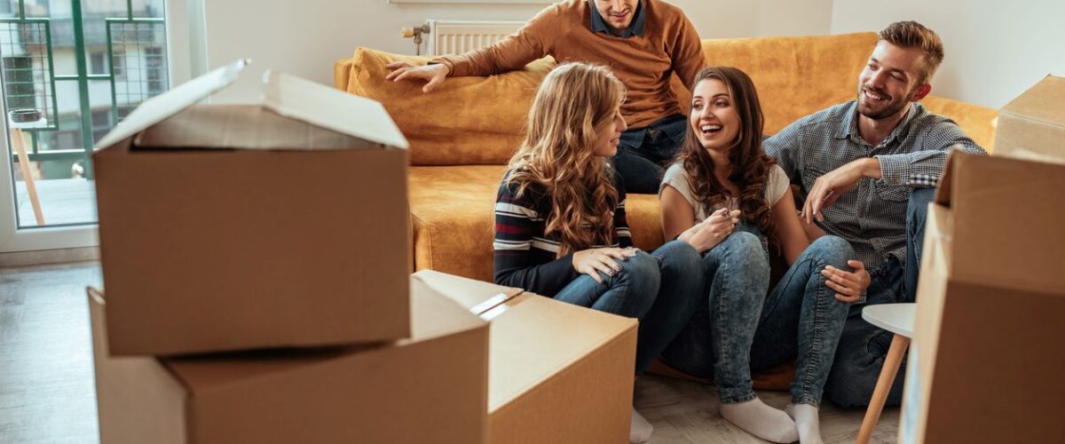 A group of roommates sharing a conversation on a couch surrounded by unpacked cardboard boxes.