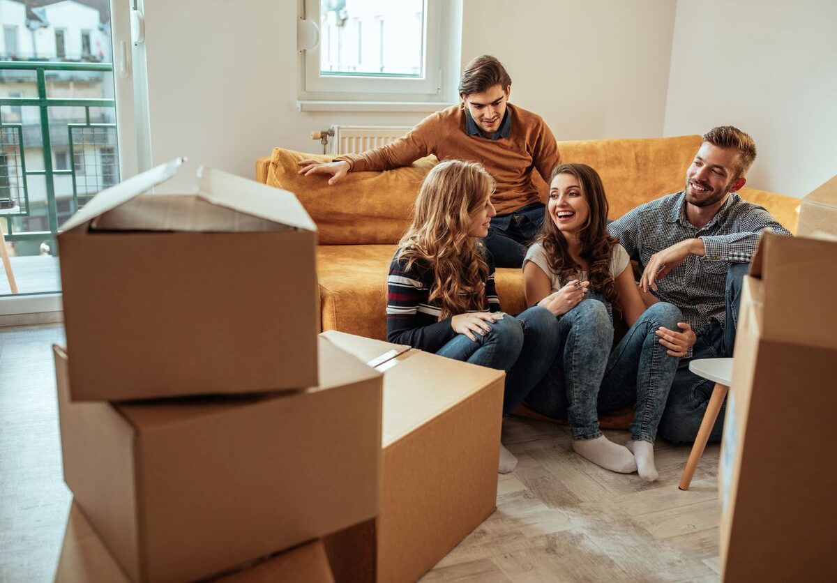 A group of roommates sharing a conversation on a couch surrounded by unpacked cardboard boxes.