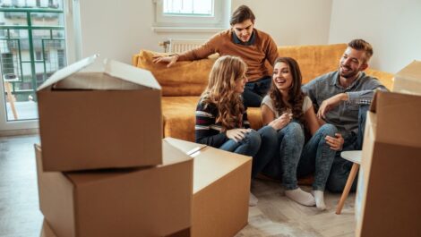 A group of roommates sharing a conversation on a couch surrounded by unpacked cardboard boxes.