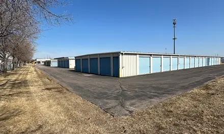 Rows of storage units beside a paved road with trees lining the area.