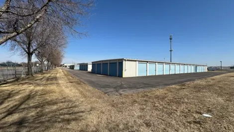 Storage units along a paved path with trees lining the side and open land.