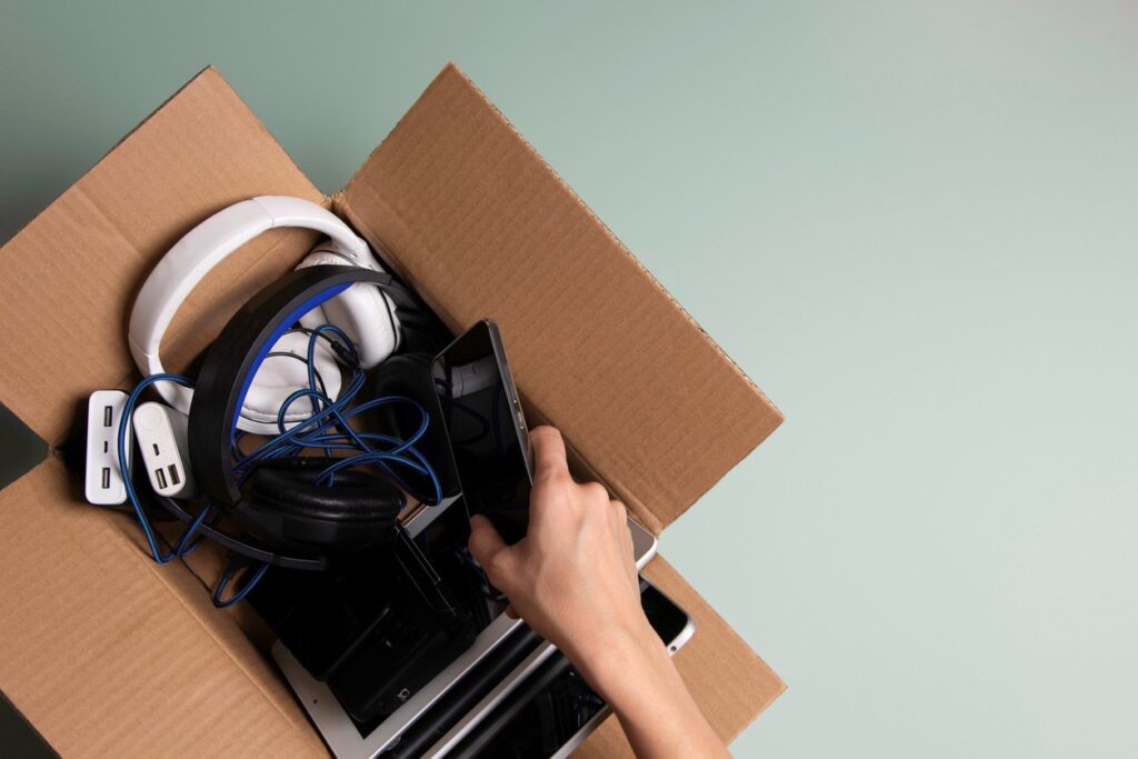 An open cardboard box filled with electronics, including headphones and cables, is being packed by hand. 