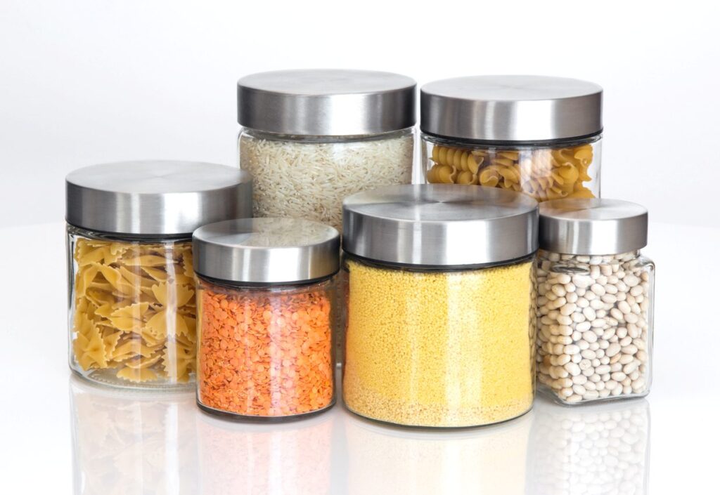 Several glass containers with silver lids, each containing dry foods, including beans, pasta, rice, and couscous.